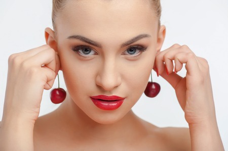 Cheerful woman is touching two cherries to her ears. She is looking at the camera with desire and temptation. Isolated on backgroundの写真素材