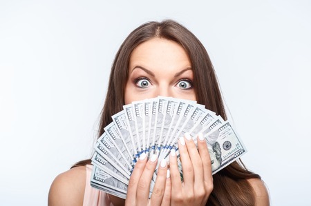 Attractive woman is holding money in her hands. She is covering her mouth with it. Her eyes are wide open with surprise. Isolated on backgroundの写真素材