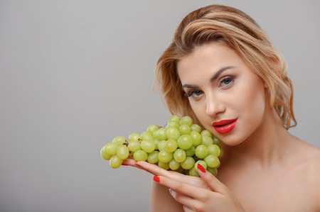 Cheerful young girl is touching bunch of grapes to her face gently. She is looking at the camera with desire. Her eyes and fingernails are red. Isolated on grey background and copy space in left sideの写真素材