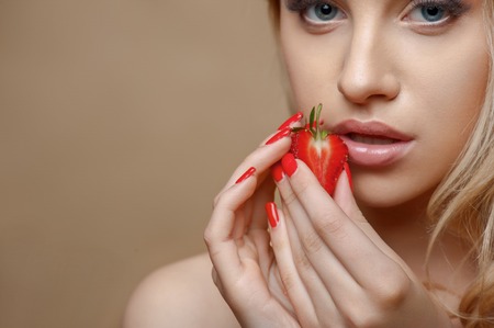 Attractive woman is holding a slice of strawberry in both her hands. She is touching it to her lips with desire. Isolated on brown background and there is copy space in left sideの写真素材
