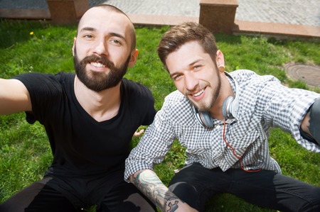 Handsome hipster guys are making selfie on grass. Their hands are stretching forward. They are...