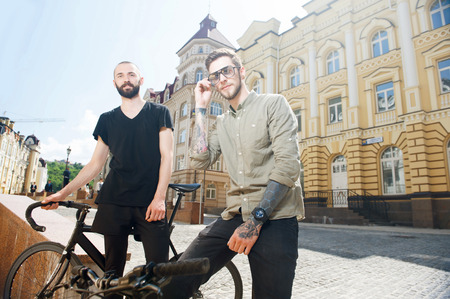 Two friends are standing near bicycles. One man is looking seriously and rearranging glasses. Another men is smilingの写真素材