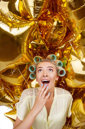 Beautiful housewife is surrounded by golden balloons. She is looking at them up with shock. She is raising her hand to wide open mouthの写真素材