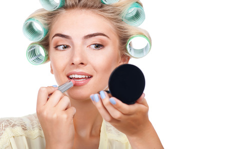 Beautiful woman is painting her lips with lipstick. She is holding mirror and looking at it with attention. The lady has curlers on her hair. Isolated on background and copy space in right sideの写真素材