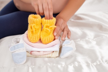 Close-up of female hands touching baby shoes with love. She is sitting on a bed near other small childlike clothingの写真素材
