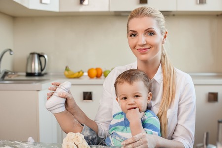 Beautiful mom is holding her son on her knees. They are preparing dough for cookies. The boy is raising his leg naughtily. The woman is touching it gently and smilingの写真素材