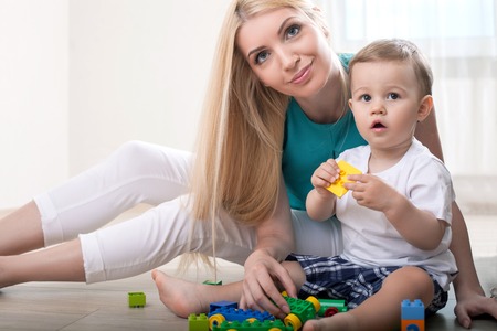 Cute mom is playing with her baby. They are holding toys and sitting on floor. The family is looking forward with interest. The woman is gently smilingの写真素材