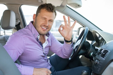 Handsome businessman is sitting at steering wheel of his car. He is showing okay sign and winking. He is smiling and looking at the cameraの写真素材