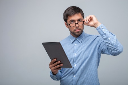 Cheerful guy is holding a tablet in his hand. He is touching his eyeglasses and looking forward...