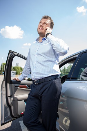Attractive businessman is talking on the mobile phone. He is standing near his car and leaning his hand on door. He is smiling and looking asideの写真素材