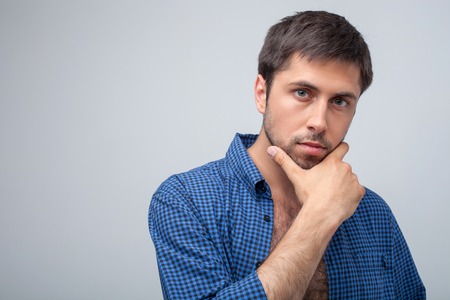 Cheerful man is looking at the camera thoughtfully. He is touching his chin seriously. Isolated on gray background and copy space in left sideの写真素材