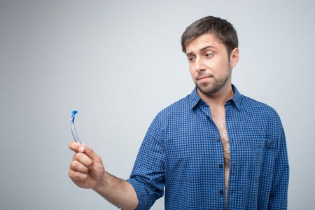 Handsome bearded guy is holding a razor in his hand. He is looking at it with doubt. Isolated on grey backgroundの写真素材
