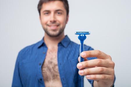 Cheerful guy is showing a razor to the camera. He is smiling proudly. Focus on a razor. Isolated on grey backgroundの写真素材