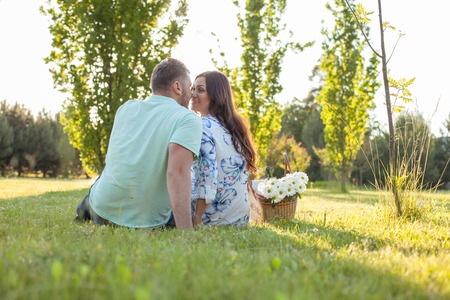 Cute loving couple is sitting on grass. They are looking at each other with love and smiling. They are touching their noses together gently. Focus on their back and copy space in right sideの写真素材