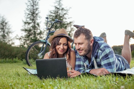 Pretty young loving couple is lying on cover in nature. The are watching film from notebook with interest. The lovers are drinking wine and smilingの写真素材