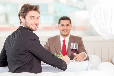 Cheerful male colleagues are sitting at the table in cafe. They are eating and laughing. The men discuss a contract and looking at the camera with joy. Copy space in right sideの写真素材