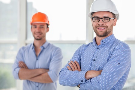 Attractive architect and handsome foreman are resting after work. They closed their hands confidently. The men are smiling and looking at the camera with joyの写真素材