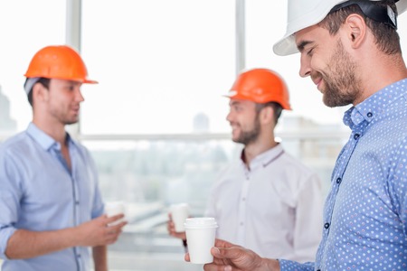 Attractive architect and his crew are drinking coffee. They are talking and laughing. Focus on the architect with white helmetの写真素材
