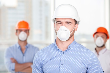 Attractive architect is standing and looking at the camera with seriousness. There are two workers in masks and helmets behind him. They are ready to work on a constructionの写真素材