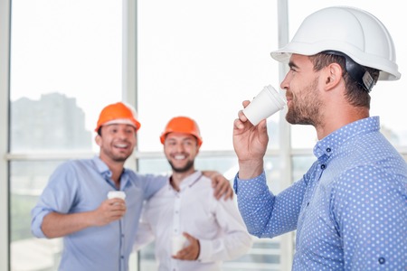 Cheerful construction team are making coffee break. The architect is drinking coffee with pleasure and smiling. Two workers are looking at him and laughing. They are embracing friendlyの写真素材