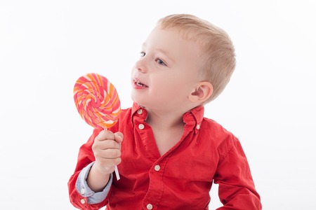 Pretty small boy is eating lollipop with pleasure. He is smiling and looking aside with joy. Isolated on backgroundの写真素材