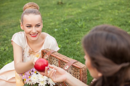 Cheerful women are sitting on green grass near a basket of food and flowers. The blond lady is giving an apple to her friend. She is looking at her with joy and smilingの写真素材