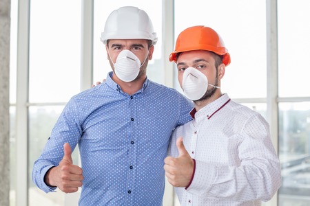 Attractive architect and cheerful foreman are working in respirators and helmets. They are giving thumbs up with joy. The men are embracing and looking at the camera happilyの写真素材