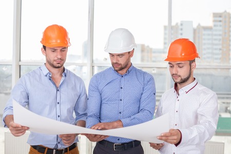 Cheerful architect is showing to builders the sketches o building. He is explaining the main ideas with seriousness. The men are looking at a paper with concentrationの写真素材