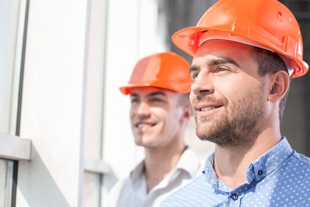 Handsome foreman and architect are standing and looking through the window dreamingly. They are smiling with hope. Their project was approvedの写真素材