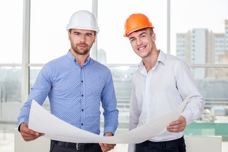Cheerful architect is explaining to foreman the plan of building. They are holding a paper of sketches. The men are looking at the camera and smilingの写真素材