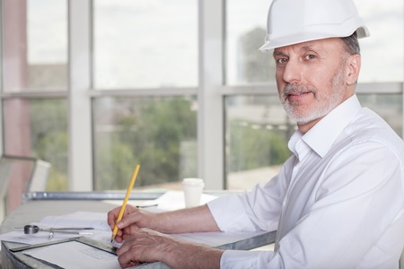 Cheerful old architect is sitting at the table and drawing sketches of building. He is looking at the camera and smilingの写真素材