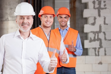 Skilled old architect and young workers are giving thumbs up. Their project was approved. The men are smiling and looking at the camera with happiness. Copy space in right side