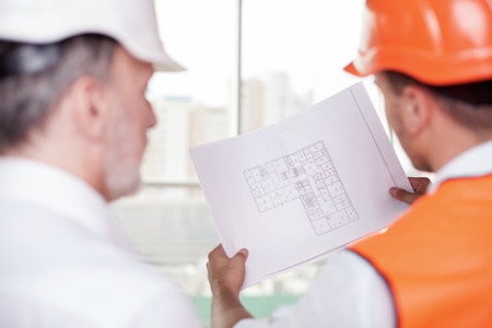 Experienced old architect and young foreman are working on plan of building. The worker is holding a blueprint. The men are looking at sketches with concentration. Focus on a paperの写真素材