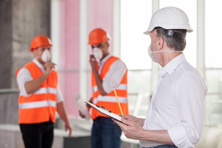 Experienced old architect is discussing with builders the concepts of building. He is writing down their ideas and looking at them with interest. The men are wearing helmets and respiratorsの写真素材