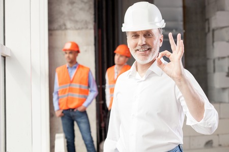 Successful senior architect is showing okay sign. He is pleased with building. The man is smiling. Handsome young workers are standing and resting behind himの写真素材