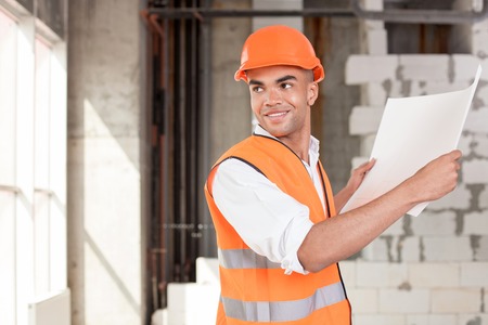 Handsome foreman is working with sketches of construction. He is holding a blueprint. The man is looking back through the window and smiling. Copy space in left sideの写真素材