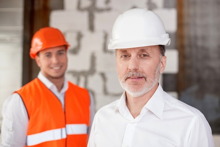 Experienced senior architect and a foreman are in process of building. They are standing and looking at the camera with joy. The men are smilingの写真素材