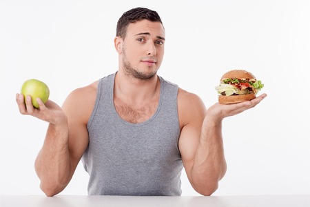 Cheerful young guy is sitting at the table. He is holding an apple and hamburger in his hands. He is puzzled. The guy is looking at the camera with hope. Isolated on backgroundの写真素材