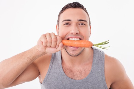 Cheerful guy is biting fresh carrot with pleasure. He is looking at the camera and smiling. Isolated on backgroundの写真素材