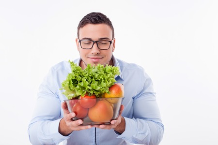 Cheerful man is holding the bowl of fruits and vegetables. He smells it with enjoyment. The man closed his eyes with pleasure. Isolated on background and copy space in right sideの写真素材