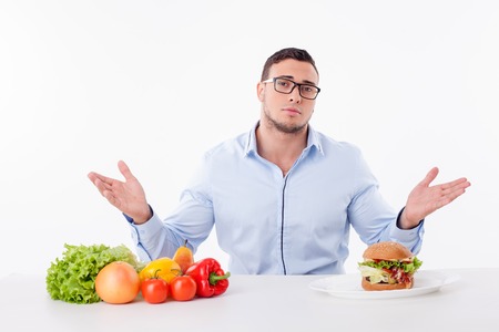 Cheerful man does not know what to choose. He is sitting at the table near healthy and unhealthy food. The man is raising his hands and asking for help sadly. Isolated on backgroundの写真素材