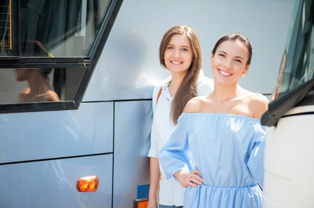 Attractive women are enjoying their trip. They are standing near a public transport. The friend are looking at the camera with joy and smilingの写真素材