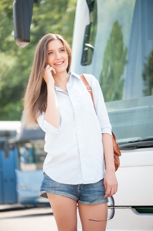 Pretty girl is talking on the mobile phone. She is standing near a bus and looking aside with joy. The lady is smilingの写真素材