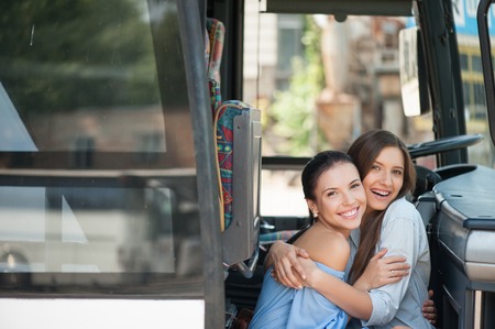 Beautiful girls are sitting on doorsteps of a bus and embracing. They are looking at the camera and smiling. Copy space in left sideの写真素材
