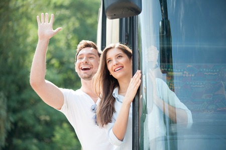 Pretty woman and man are standing in doors of the bus. They are looking through it and laughing. The boy is raising his palm and helloing with everybodyの写真素材