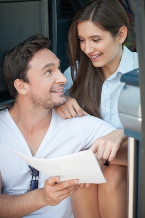 Cheerful young man and woman begin their trip. They are sitting on steps of  bus and smiling. The man is showing to his girlfriend a route of their travel. They are looking at each other and embracingの写真素材