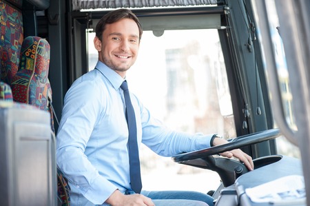 Handsome bus driver is sitting at steering wheel. He is looking at the camera and smiling