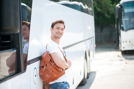 Handsome man is standing near a bus and leaning on it. He is holding a rucksack and smiling. The man is looking at the camera with joy. Copy space in right sideの写真素材