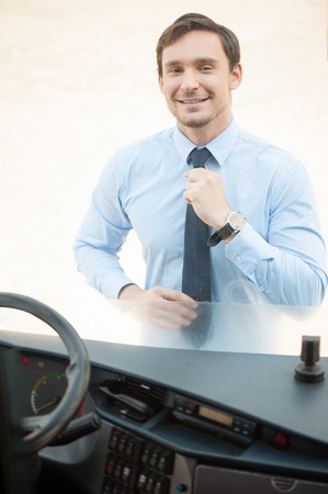 Cheerful male driver is standing outside. He is looking through the window of a bus and adjusting his tie. The guy is smilingの写真素材