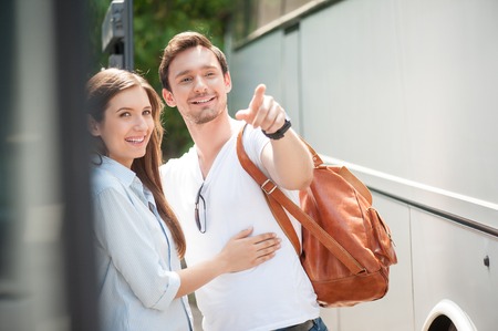Cute loving couple is spending time together near a public transport. They are enjoying their journey. They are embracing and smiling. The guy is pointing finger forward with joyの写真素材